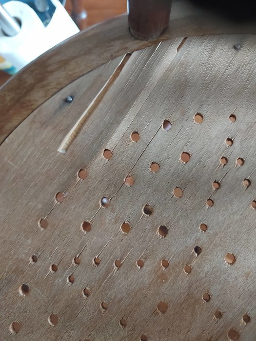 Närbild av sliten stolsits i plywood med hålmönster och synbara skador.