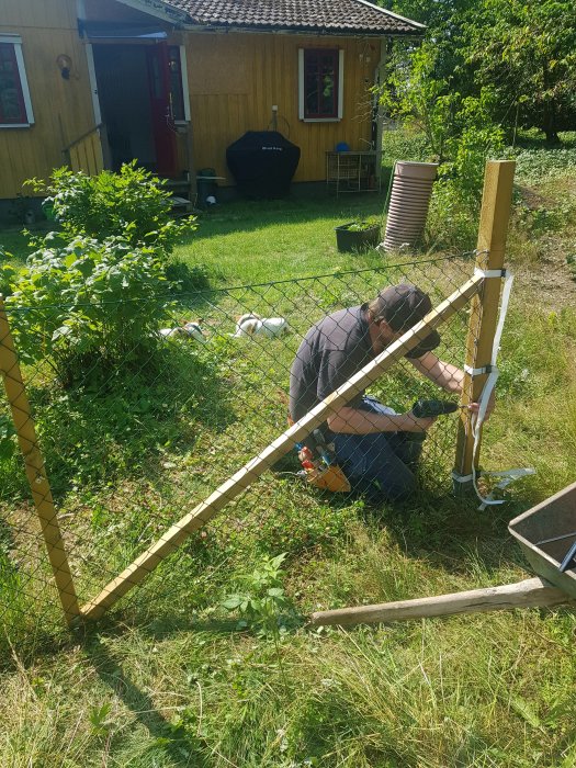 Person som bygger staket i en trädgård med grönska och ett hus i bakgrunden.