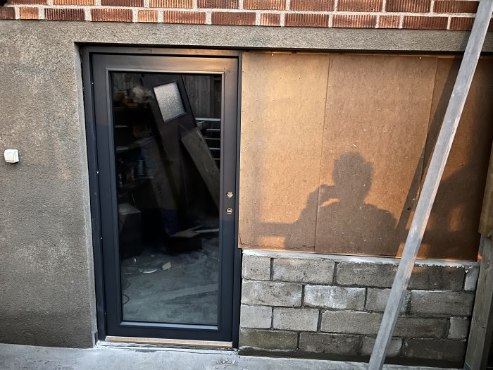 En ytterdörr med delvis reflekterande glas och en shadow på en MDF-panel bredvid där ett fönster är planerat.