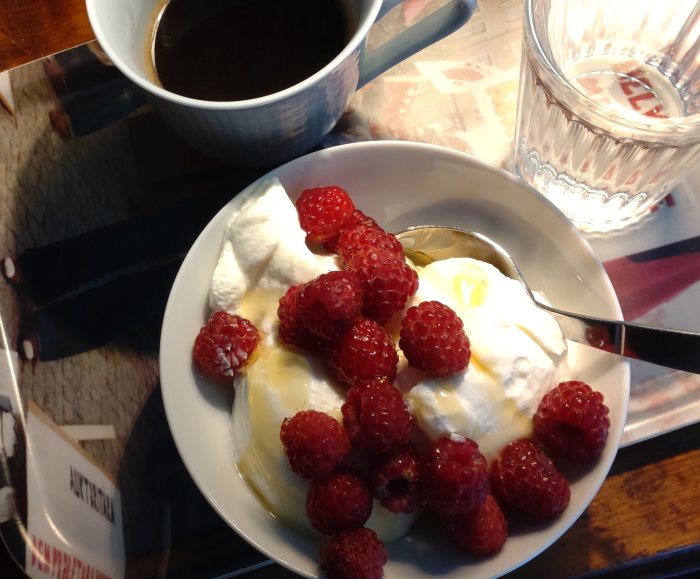 En skål med yoghurt och hallon bredvid en kopp kaffe och ett glas vatten på ett bord.