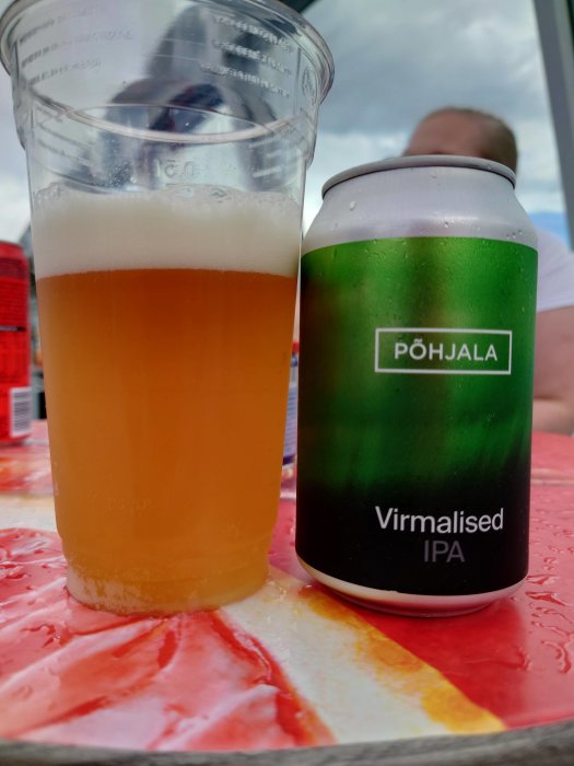 En burk Põhjala Virmalised IPA bredvid ett plastglas fyllt med öl på ett färgglatt bord.