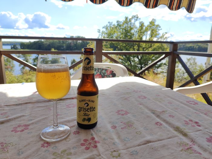 Flaska och glas med veteöl på ett blommigt bord utomhus med sjö och träd i bakgrunden.