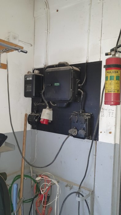 Eldistributionssystem med säkringsskåp och rörledningar på en garagevägg, brandsläckare till höger.