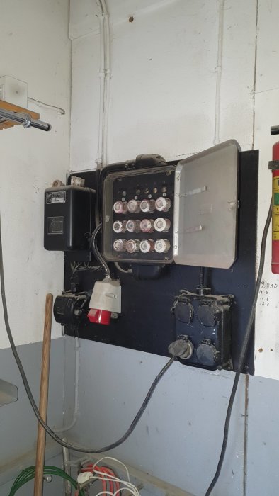 Gammalt proppskåp och elmätare på garagevägg, elinstallationer syns med säkringar och ledningar.