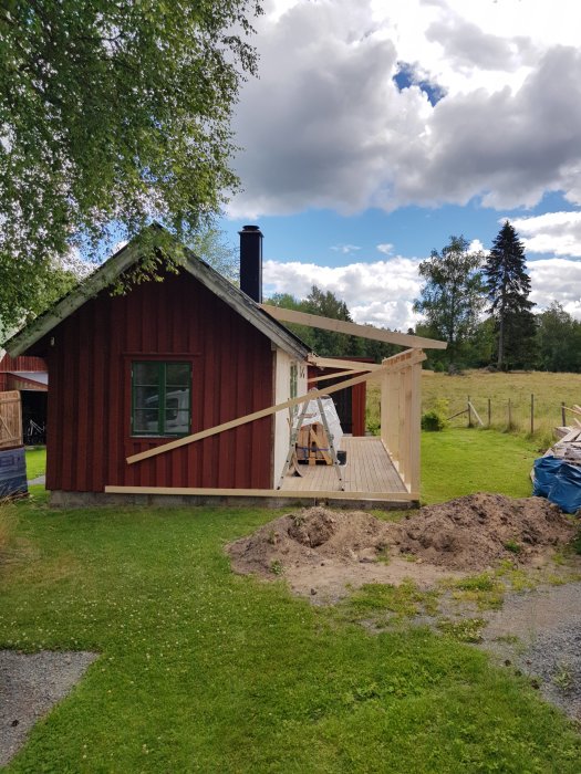 Röd stuga under renovering med nybyggd träställning för takarbete, omgiven av grönt gräs och träd.