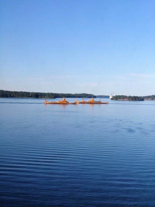 Båtar på lugnt vatten med skogklädd kustlinje i bakgrunden och klarblå himmel.