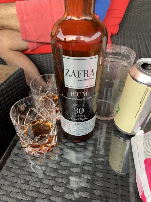 Flaska Zafra 30-årig rom och ett glas med rom på ett vävt bord, bakgrund med personer.