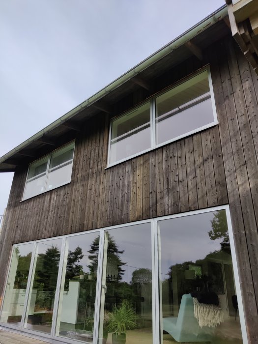 Ett tvåvåningshus med träfasad och stora fönster visar tecken på väderpåverkan och solblekning på panelerna.