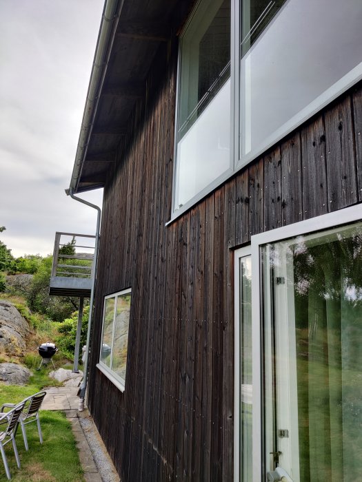 Fasaden på ett hus med mörk träpanel och synliga galvade spikar, stora fönster och en balkong, omgiven av natur.