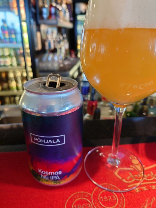 Öppnad ölburk märkt 'Põhjala Kosmos NE IPA' bredvid ett glas fyllt med skummande öl på en bar med dryckesflaskor i bakgrunden.