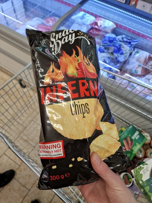 Hand håller en påse Snack Day Inferno chips med varningsmärkning "EXTREMELY HOT" i en matvarukorg.