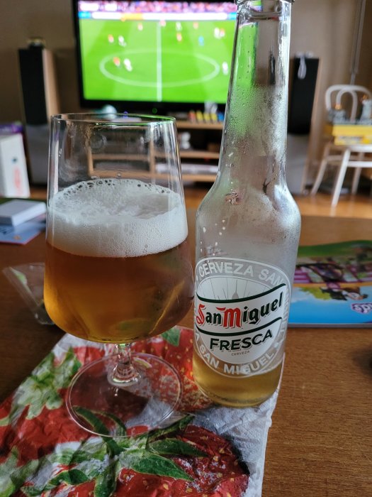 Öppnad San Miguel Fresca-flaska bredvid ett glas öl, med fotbollsmatch i bakgrunden.