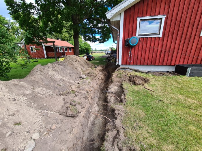 Nygrävd ränna för vattenledning nära ett rött hus med en synlig vattenslang och jordhögar.