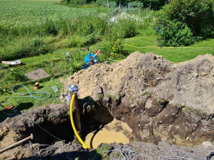 Nygrävd brunn med gult rör i ett grävt dike omringat av gräshögar och verktyg.