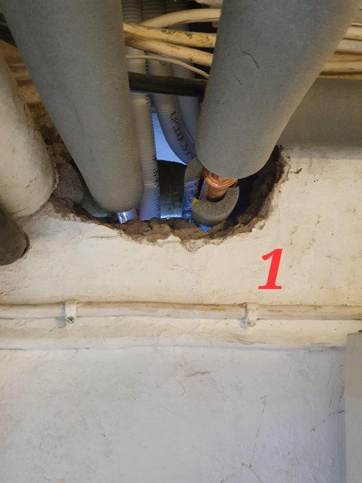 Avloppsrör och flexibla pexrör som sträcker sig genom ett hål i en betongvägg märkt med rött nummer 1.