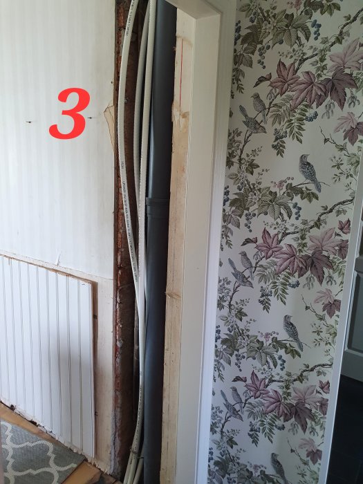 Avloppsrör och pexrör som hänger fritt vid en öppen väggsektion, vid tapeterat rum.