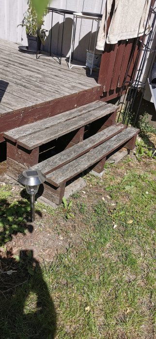 Sliten trätrappa med tre steg leder upp till en altan, med tydliga tecken på åldring och väderpåverkan.