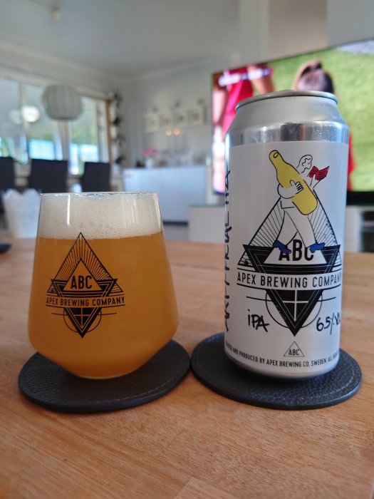 Ett glas öl och en burk från Apex Brewing Company på ett bord med en suddig inredningsbakgrund.