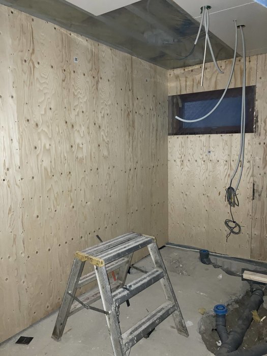 Ett renoverat badrum med isolerade väggar av plywood, avloppsrör på golvet, och en stege i förgrunden.