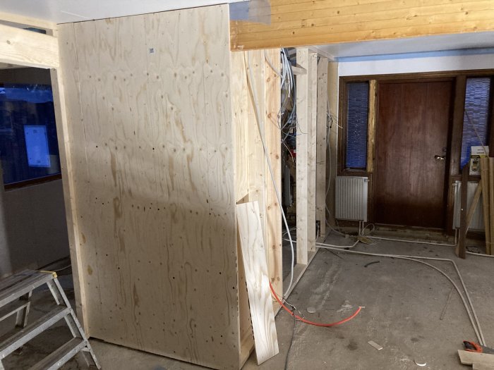 Rensat rum med plywoodvägg, takbjälkar och kablar, redo för renovering.