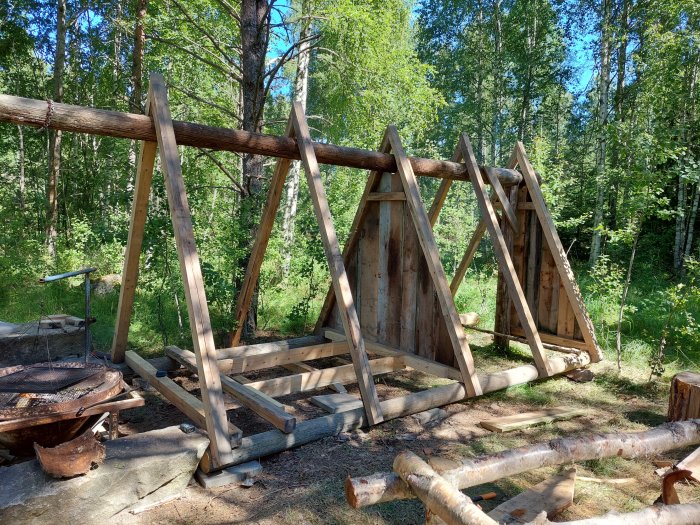 Pågående bygge av vindskydd vid grillplats i skogsmiljö, med träreglage och presenning på golvet.