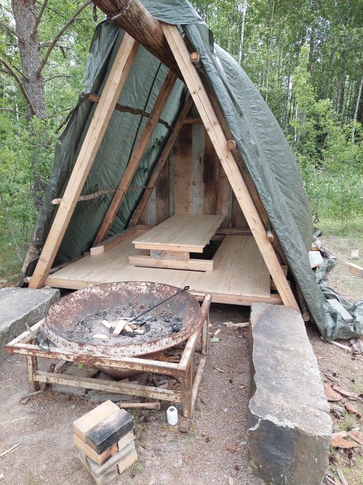 Utomhus grillplats under konstruktion med vindskydd, uppfällbart bord och presenningståtak i skogsmiljö.