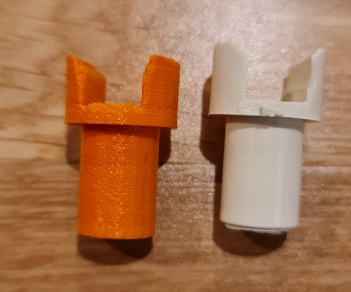 Två objekt utskrivna med 3D-skrivare, ett i orange och ett i vitt, som ligger på en träyta.