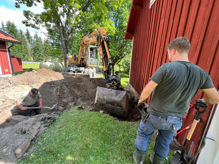 Grävningsarbete vid torp med grävmaskin och två personer på en tidigare fin gräsmatta.