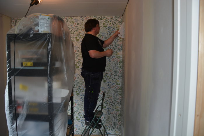 Person i arbetskläder tapetserar en vägg med blommig tapet i ett rum under renovering.