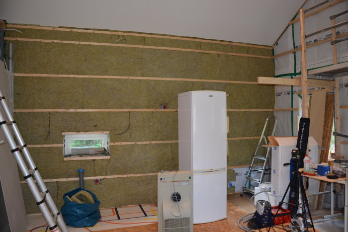 Renoveringsprojekt med isolering och träreglar på en vägg, en stege, och renoveringsmaterial i ett rum under uppbyggnad.