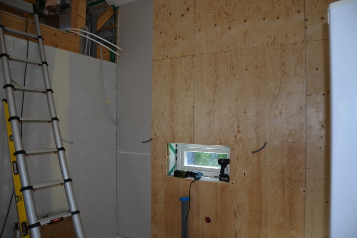 Renoveringsarbete med halvfärdig vägg av OSB-skivor, gipsskiva och isolering nära ett litet fönster och en stege.