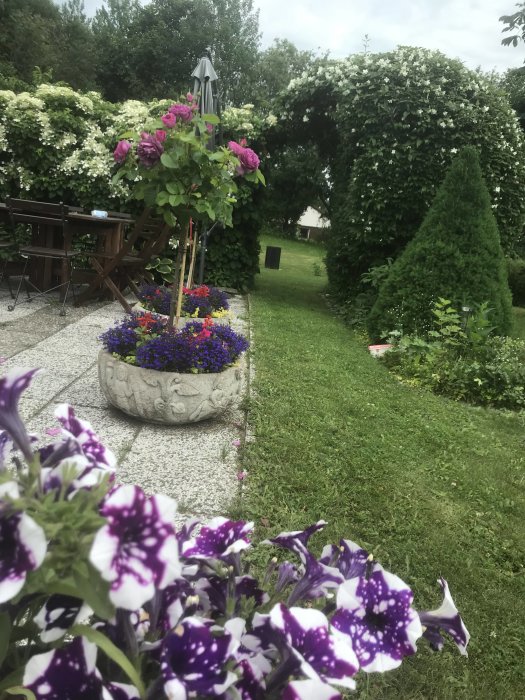 Trädgård med blomsterarrangemang och två ihopfällbara bord under blommande buskbonad.