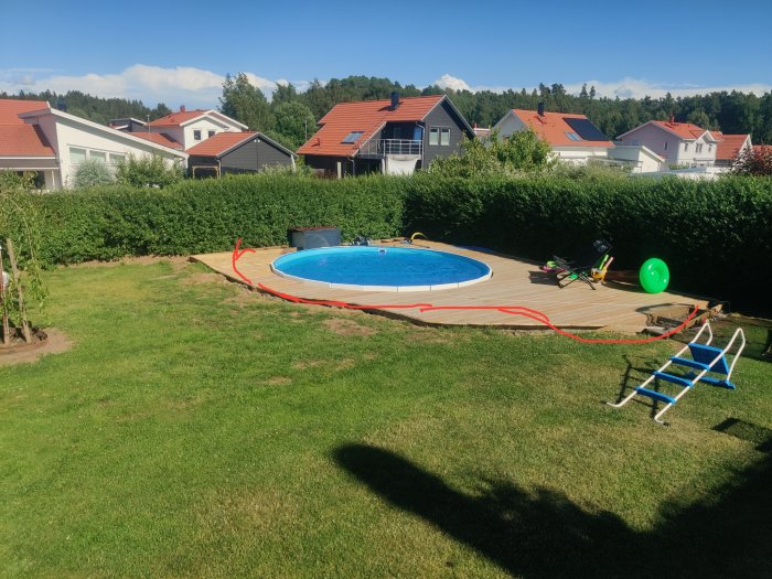 Trädgård med rund pool, trätrall, röd markering längs gräskant, leksaker och hus i bakgrunden.