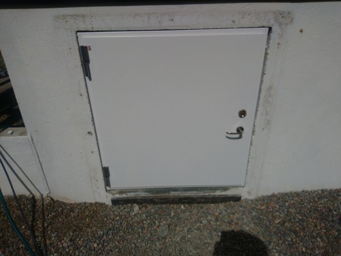 En vit dörr med lås och handtag monterad i en vitmålad vägg, med en tröskel av grus.