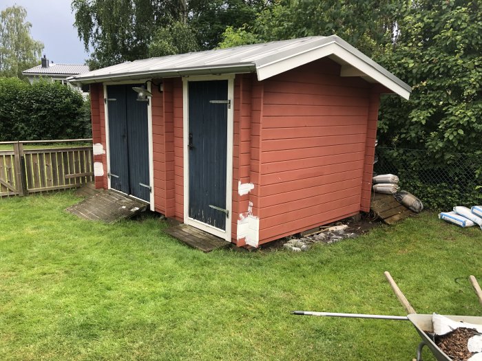 Röd träfriggebod med blå dörrar och vita omålade områden redo för målning, omgiven av grönt gräs.