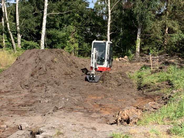 Tvåtons grävmaskin på en grävplats omsluten av träd, med stora jordhögar i bakgrunden.