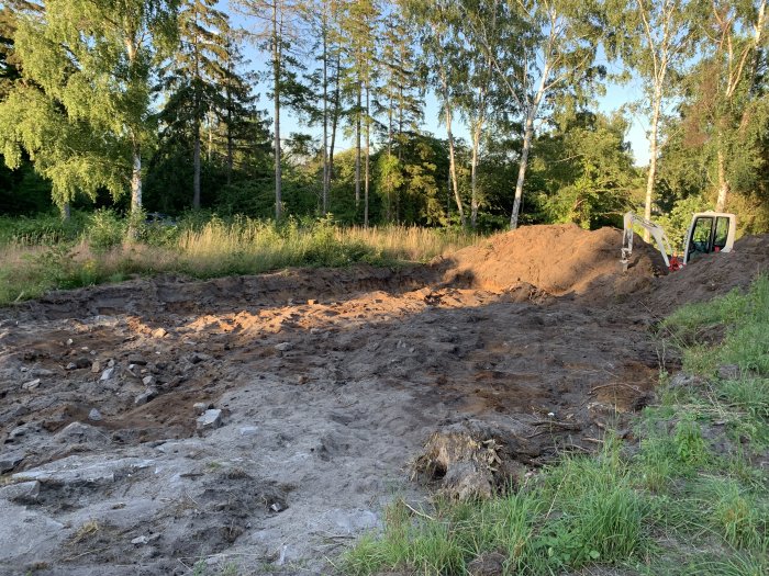 Tvåtons grävare vid utgrävd grund med jordhögar på en byggplats omgivet av träd.