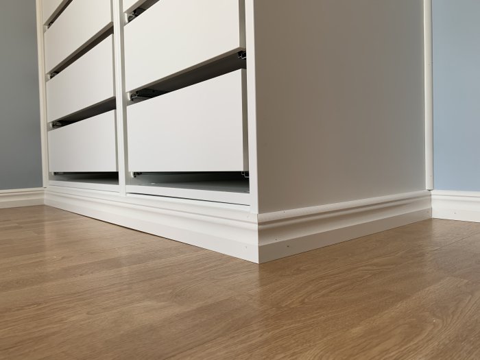 Nylagda vita golvsocklar i hörnet av ett rum med ljusbrunt laminatgolv och en vit byrå.