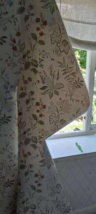 Ny tapet med blom- och växtmönster i ett sovrum, delvis uppe vid ett hörnfönster med verktyg synligt.