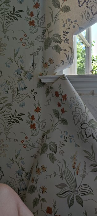 Blommig tapet med varierande gröna, blåa och orange blommönster ojämnt monterad vid ett fönster med synlig veckning.