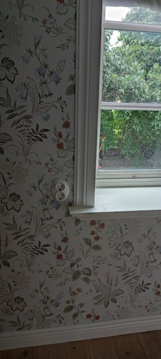 Ny tapetsering i sovrum med mönstrad tapet vid fönster och eluttag på väggen.