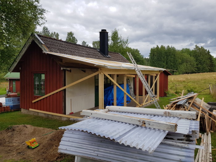 Röd stuga under renovering med ny råspont på taket, byggmaterial framför och leksakstraktor i en jordhög.