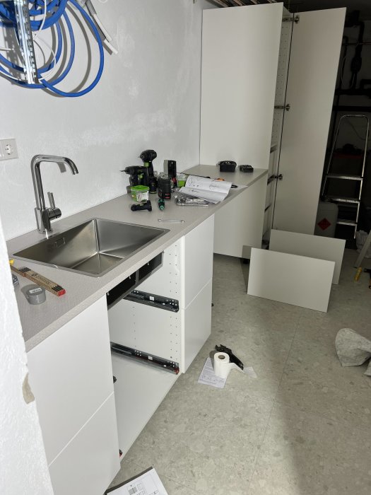 Ej färdigställd tvättstuga med öppna skåp och installation av diskho, verktyg och ritningar utspridda.