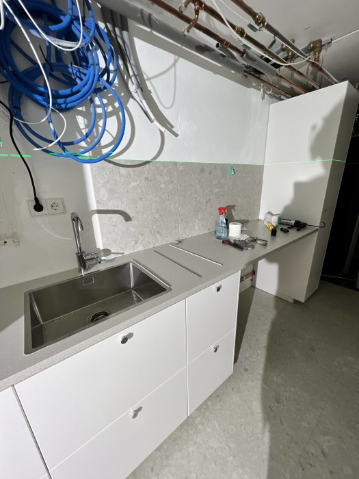 Tvättstugan under renovering med vit skåpinredning och rostfri diskho, omgiven av kablar och rör.