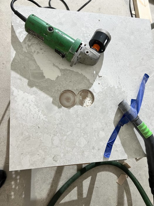 Håltagning i stenbänkskiva med grön elektrisk borr och diamantkärna, dammsugarslang och blå tejp i bakgrunden.