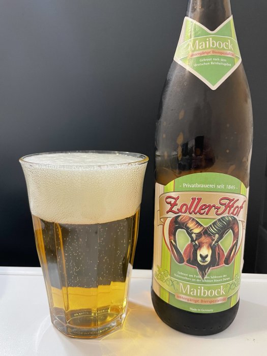 Ölglas med skum vid sidan av en flaska Maibock från Zoller Hof på en vit yta.