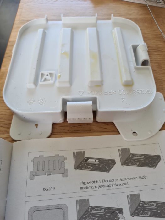 Vit bottenpanelskydd för tvättmaskin och monteringsinstruktioner på ett bord.