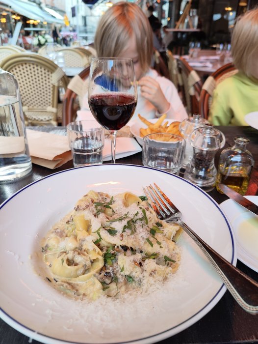 Tallrik med tryffelpappardelle framför ett glas rödvin, med barn som äter i bakgrunden på en uteservering.