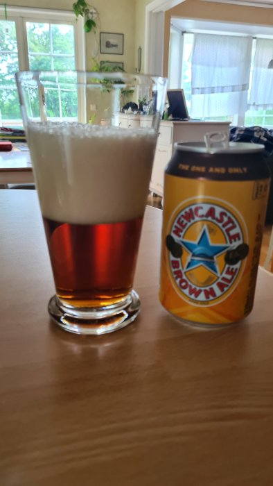 Ett glas fyllt med öl bredvid en burk Newcastle Brown Ale på ett bord inomhus.