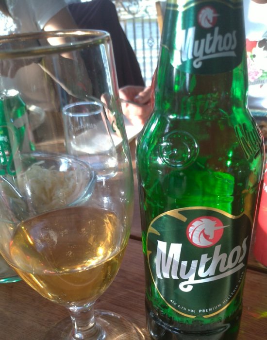 En halvtom ölflaska av märket Mythos bredvid ett glass ölglas på ett bord.